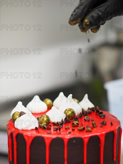 Chef sprinkling gold dust chocolate over a frosted dripping red brown dark chocolate cake with meringue decorations