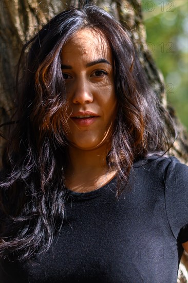 Smiling Cheerful hispanic young woman in black shirt standing in bright sunlight by a tree, blurred background with bokeh, daytime, AI generated