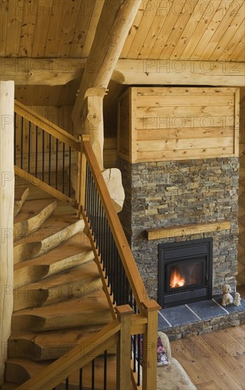 High angle view of wooden and black wrought iron staircase and lit natural stone fireplace in living room inside handcrafted Eastern white pine Scandinavian log cabin home, Quebec, Canada, North America