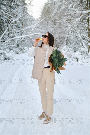 Playful woman holding fir branches under her arm standing on winter snowy road and drinking champagne
