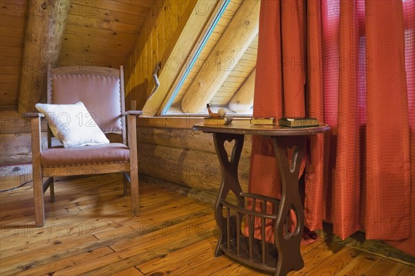 Brown wooden leather armchair with white cushion and side table in master bedroom with red curtains and wood plank floor on upstairs floor inside rustic contemporary Scandinavian log cabin home, Quebec, Canada, North America
