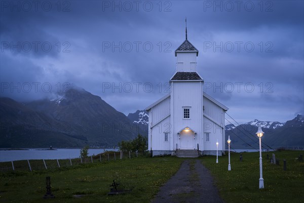 The white church of Gimsoy (Gimsoysand church) by the sea. A path leads to the church. The church and lanterns are illuminated. At night at the time of the midnight sun. Cloudy sky. Early summer. Gimsoya, Lofoten, Norway, Europe
