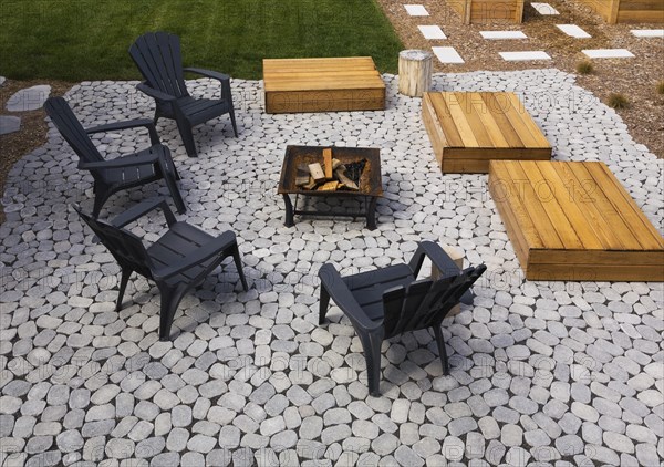 Grey paving stone patio with black Adirondack sitting chairs, slightly raised flat rectangular red cedar wooden bench platforms around a metal firepit in residential backyard in summer, Quebec, Canada, North America