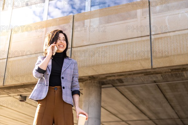 Young smart casual latin businesswoman walking down the street talking on the phone holding glass of coffee. Smiling girl in jacket talking on the phone in the street, holding a coffee