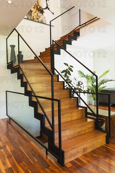 Black powder coated steel L-shaped staircase with stained and varnished wooden steps and tempered glass handrail in living room with acacia wood flooring inside contemporary home, Quebec, Canada, North America