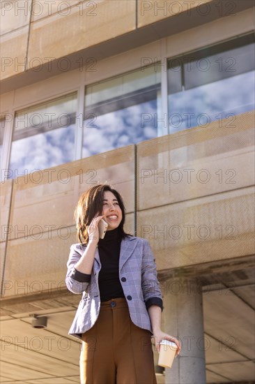 Young smart casual latin businesswoman walking down the street talking on the phone holding glass of coffee. Smiling girl in jacket talking on the phone in the street, holding a coffee