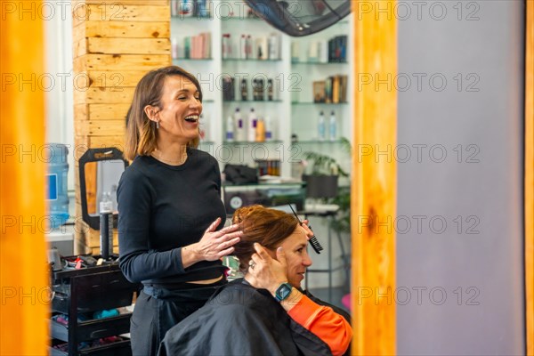 Reflection view on a mirror of a hairdresser laughing while talking and combing a female customer in the salon