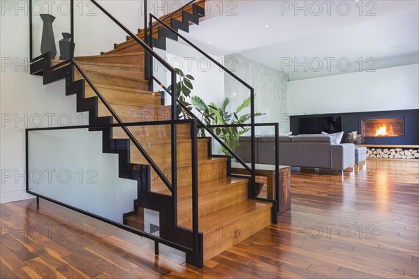 Black powder coated steel L-shaped staircase with stained and varnished wooden steps and tempered glass handrail in living room with grey leather L-shaped sectional sofa and acacia wood flooring inside contemporary home, Quebec, Canada, North America