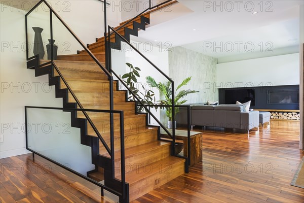 Black powder coated steel L-shaped staircase with stained and varnished wooden steps and tempered glass handrail in living room with grey leather L-shaped sectional sofa and acacia wood flooring inside contemporary home, Quebec, Canada, North America