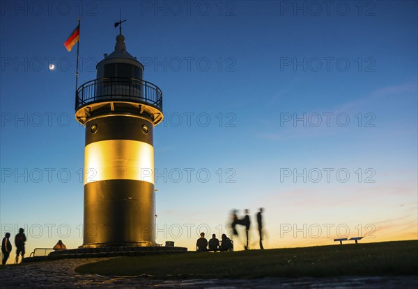 Black and white lighthouse, called 'Kleiner Preusse', stands illuminated at night shortly after sunset at the end of a paved, curved path, silhouettes of people next to it, wipe effect, path in the foreground, the waxing moon is in the blue sky, Crescent with visible dark moon disc, Germany flag, evening, close-up, North Sea resort of Wremen, Lower Saxony Wadden Sea National Park, World Heritage Site, Wurster North Sea coast, Land Wursten, Cuxhaven district, Lower Saxony, Germany, Europe