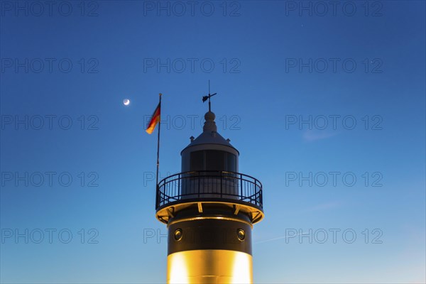 Black and white lighthouse, called 'Kleiner Preusse', stands illuminated at night shortly after sunset in front of the blue sky, the waxing moon shines as a crescent with visible dark moon disc, Germany flag, close-up, close-up, North Sea resort Wremen, Lower Saxony Wadden Sea National Park, World Heritage Site, Wurster North Sea coast, Land Wursten, district of Cuxhaven, Lower Saxony, Germany, Europe