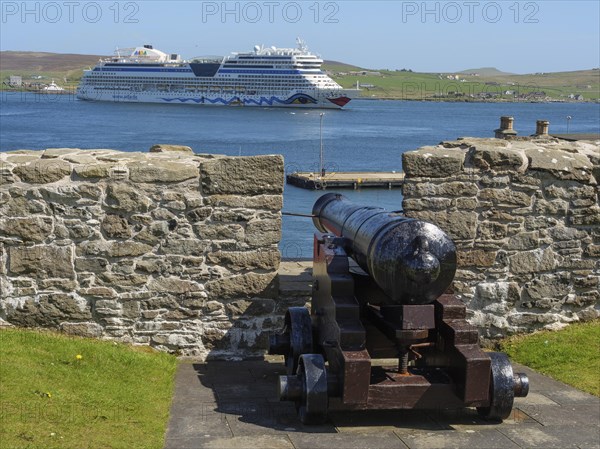 A cannon aims at a large ship in the sea behind a wall, old cannons on a stone wall by the sea against a blue sky, Lerwick, Scotland, United Kingdom, Europe