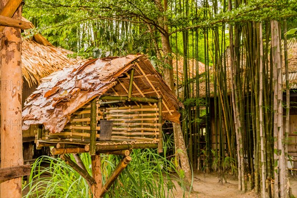 Thai spirit house mounted on a pillar with buildings and bamboo trees in background in Thailand