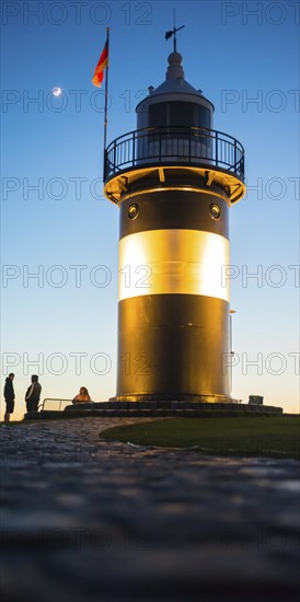 Black and white lighthouse, called 'Kleiner Preusse', stands illuminated at night shortly after sunset at the end of a cobbled, curved path, silhouettes of people in the background, path in the foreground, in the blue sky the waxing moon shines as a crescent with visible dark moon disc, Germany flag, close-up, North Sea resort Wremen, Lower Saxony Wadden Sea National Park, World Heritage Site, Wurster North Sea coast, Land Wursten, district of Cuxhaven, Lower Saxony, Germany, Europe