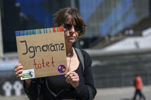 Participant in the silent protest for the hunger strikers. Her placard reads: Ignorance kills . Stickers from Mothers Rebellion and Extinction Rebellion. In the background, crosses for those who died at the Wall. Paul Loebe House, Berlin, 12 May 2024