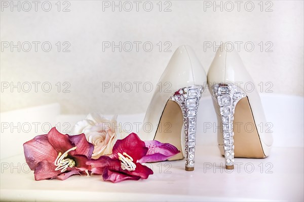 Fancy white high heels with rhinestone details next to pink flowers on a white bench creating an elegant bridal atmosphere