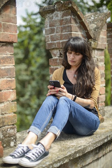 Young woman in casual clothing focused on conversation with smartphone in a peaceful garden with blurred brickwork in the foreground