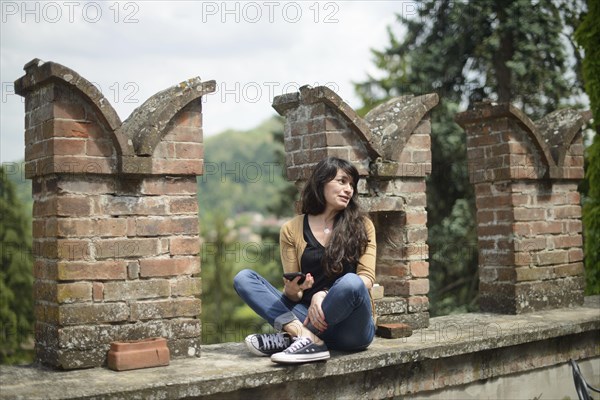 Young woman in casual clothing focused on conversation with smartphone in a peaceful garden with blurred brickwork in the foreground