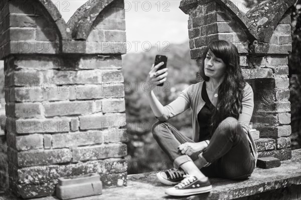 Young hispanic woman in casual clothing focused on conversation with smartphone in a peaceful garden with blurred brickwork in the foreground