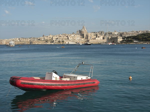 A red inflatable boat floats calmly on the sea in front of the city with historic buildings in the background, Valetta, Malta, Europe