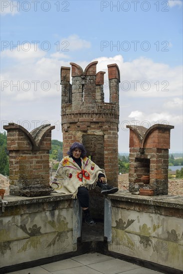 Contemplative older woman sits on a brick balcony overlooking a lush landscape, her expression serene, wearing a floral shawl