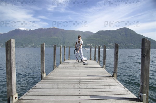 Happy and Elegant Woman Cleaning with a Vacuum Cleaner on a Pier over an Alpine Lake Maggiore with Mountain in a Sunny Day in Brissago, Ticino, Switzerland, Europe