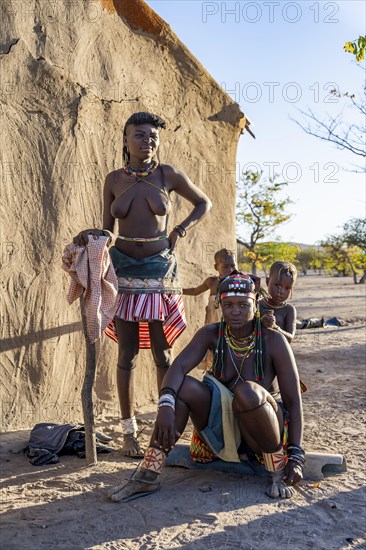 Two Hakaona woman with traditional kapapo hairstyle, with children, in front of their mud hut, in the morning light, in the morning light, Angolan tribe of the Hakaona, near Opuwo, Kunene, Namibia, Africa