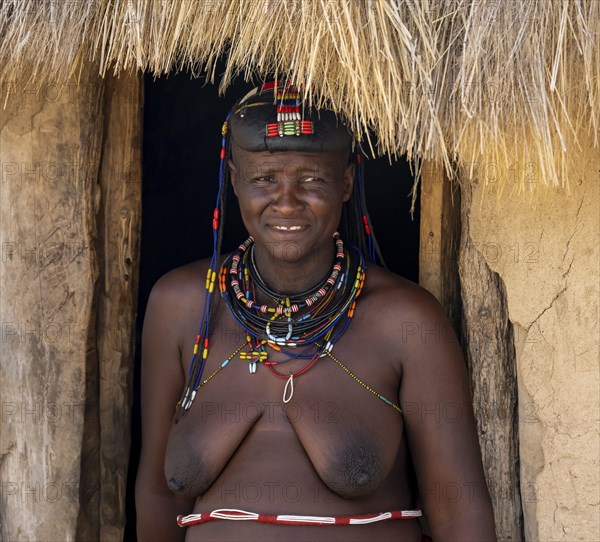 Hakaona woman with traditional kapapo hairstyle, in the entrance of her mud hut, portrait, in the morning light, Angolan tribe of the Hakaona, near Opuwo, Kunene, Namibia, Africa