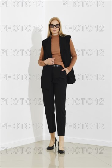 Serious woman in black vest, terracotta turtleneck, and breeches standing against a white office wall