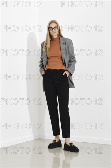 Stylish woman wearing checkered blazer and brown turtle neck sweater and black trousers standing against white office wall