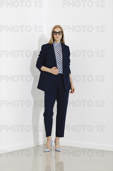 Professional woman stands confidently against a white wall, dressed in a chic navy blue business suit paired with light blue heels