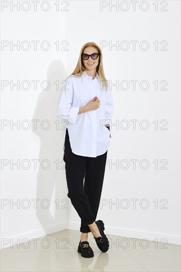 Urban-style fashion portrait of happy smiling woman wearing white shirt and black trousers. Cheerful woman standing with crossed legs in a bright room