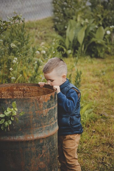 A young boy in a blue jacket and brown pants peers into a rusted metal barrel in a grassy outdoor setting.Belarus.Minsk