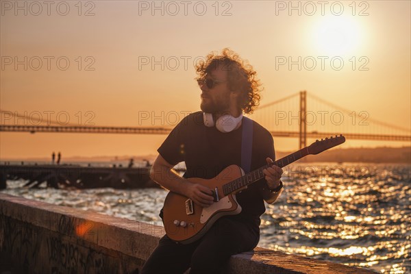 Hipster street rock musician in black playing electric guitar in the street on sunset on embankment with 25th of April bridge in background. Lisbon, Portugal, Europe