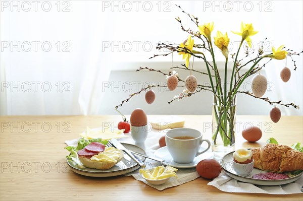 Easter breakfast served on a wooden table with a bouquet from daffodil flowers, spring branches and hanging egg decoration, copy space, selected focus, narrow depth of field