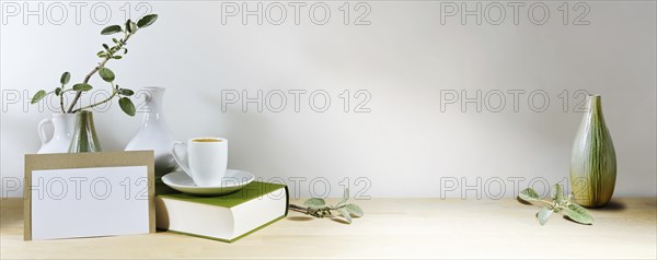 Mock-up of a blank greeting card, vases with sage leaf branch and coffee cup on a book, wooden table or desk against a gray wall, home office arrangement, panoramic format, copy space, selected focus