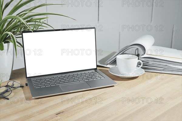 Mock up on an open laptop with a blank white screen, coffee cup and ring binder on a wooden office desk against a white painted wall, business concept, copy space, selected focus