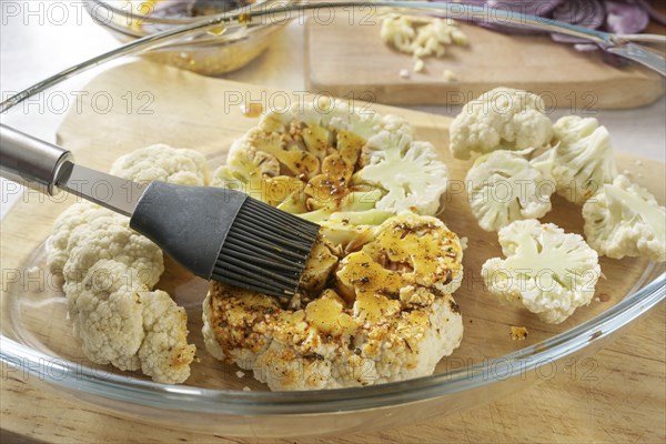 Raw cauliflower slices in a glass casserole are coated with a mixture of spices, herbs and olive oil by a silicone brush, cooking preparation for baked vegetable steaks, selected focus, narrow depth of field