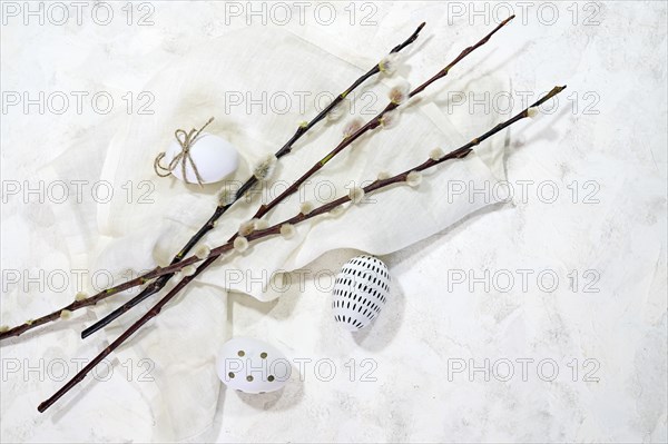 White Easter eggs with small black decor and some willow branches with catkins on a light cloth and background, copy space, high angel view from above, selected focus