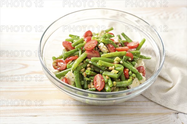 Green bean salad with tomatoes, pine nuts and feta cheese in a glass bowl on a light wooden table, Mediterranean snack or antipasto, copy space, selected focus, narrow depth of field