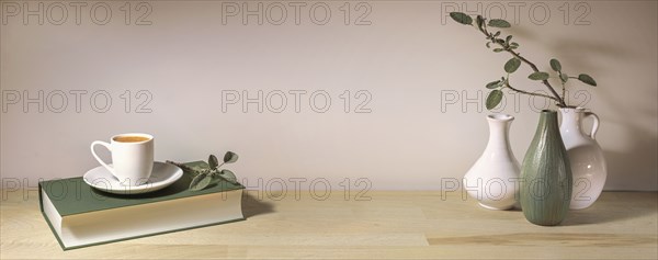 Coffee cup on a gray green book and a group of vases with a sage leaf branch on a wooden table table against a pale wall, panoramic format, home decor, copy space, selected focus