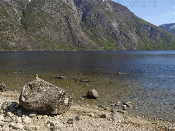 A large boulder lies on a rocky beach with a tranquil fjord and mountains beyond, Eidfjörd, Norway, Europe