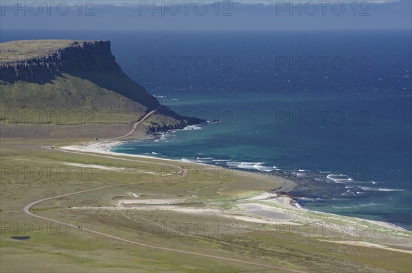 A cliff juts out over the deep blue sea, next to a long, sandy beach and green meadows, Latrabjarg, Vestfirdir Peninsula, Iceland, Europe