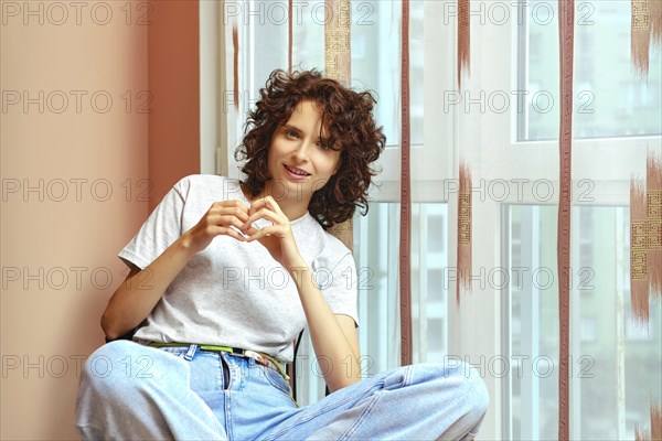 Cheerful young woman with curly hair sitting by window and smiling