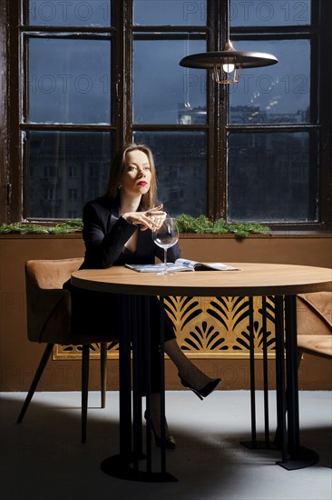 Lonely sad woman in pantsuit sitting behind empty table in cafe and reading magazine