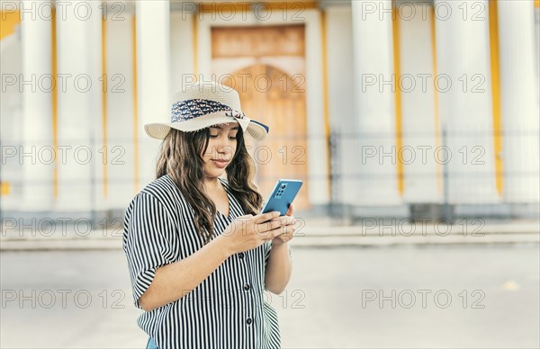 The girl wears a hat, stands in front of a Catholic church with a blurred background, is smiling and sending messages from her cell phone