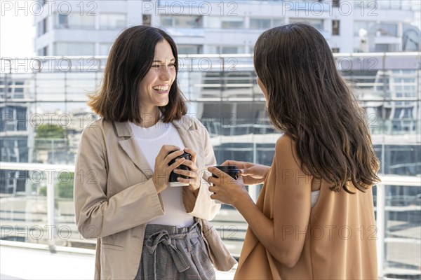 Two businesswomen are standing outside a building, enjoying their office break. They are smiling and holding cups of coffee, indicating a sense of camaraderie and friendship