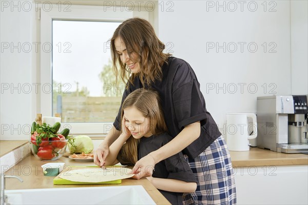 Mother and daughter spread cream cheese on a flatbread together while standing in a bright kitchen early in the morning