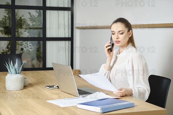 Businesswoman sitting at a desk in office, holding a document and speaking on the phone with customer