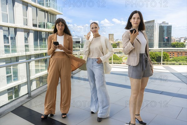 Three businesswomen holding smartphones are looking at the camera while posing for the photo